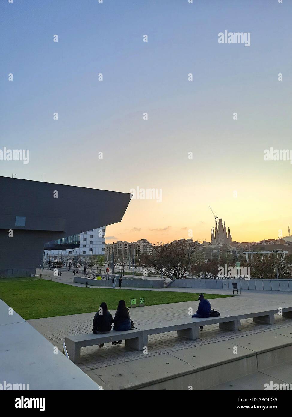 Espace public contemporain avec des bancs et une pelouse verte où les gens s'assoient en profitant de la vue du coucher du soleil sur la Sagrada Familia au loin Banque D'Images