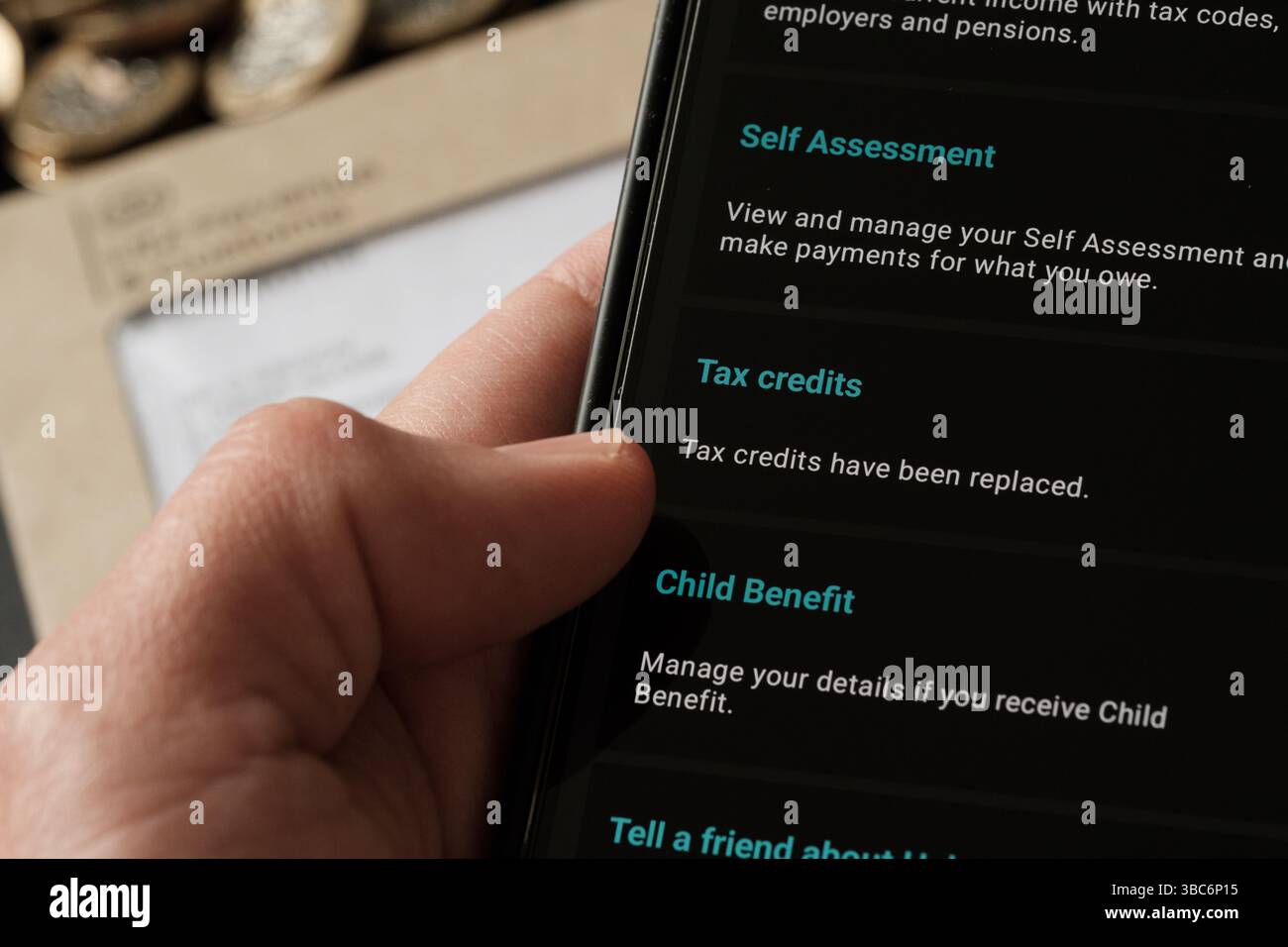 Personne utilisant l'application mobile HMRC pour voir l'auto-évaluation, les crédits d'impôt et les services de prestation pour enfants, avec l'enveloppe du gouvernement britannique et les pièces en arrière-plan, Sta Banque D'Images