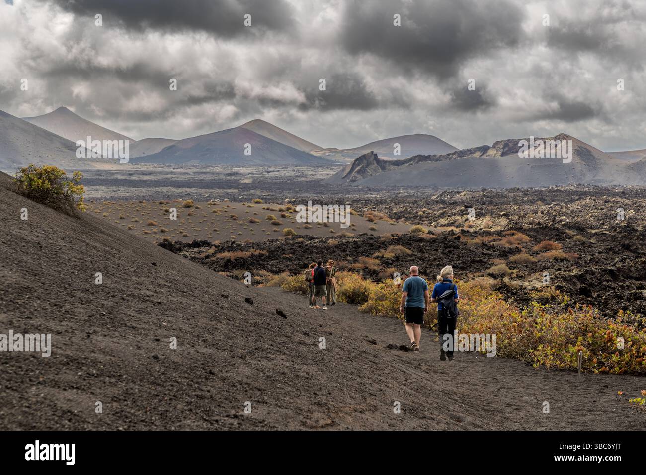 Un groupe de randonneurs marche le long d'un sentier sombre de lapilli volcaniques à travers la zone volcanique près de Mancha Blanca à Lanzarote. Plusieurs cônes volcaniques du Parque Natural de los Volcanes s'alignent en arrière-plan. Des arbustes verts poussent dispersés le long du chemin. Îles Canaries, Espagne. Banque D'Images