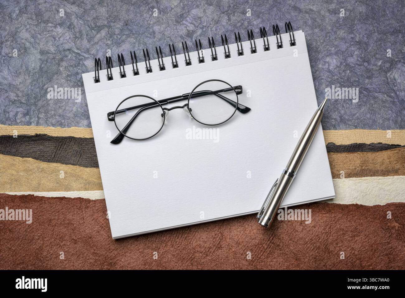 Carnet de croquis d'art en spirale vierge avec un stylo et des lunettes de lecture, lunettes de lecture contre le paysage de papier abstrait dans des tons de terre Banque D'Images