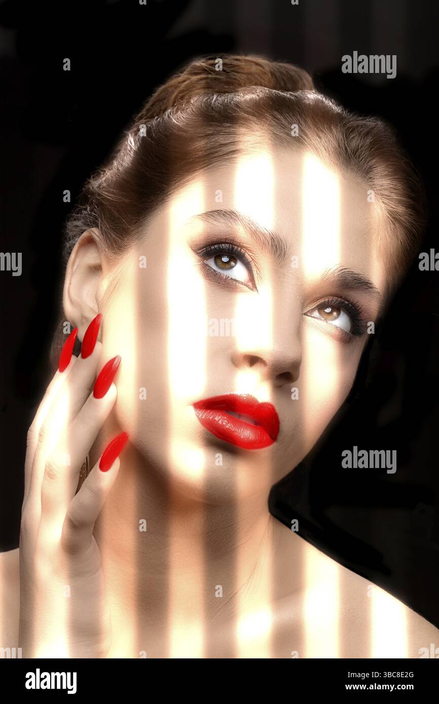 Un modèle avec rouge à lèvres rouge et vernis à ongles est illuminé par la lumière vive des stores sur un fond sombre. Le visage d'un beau, bien-groome Banque D'Images