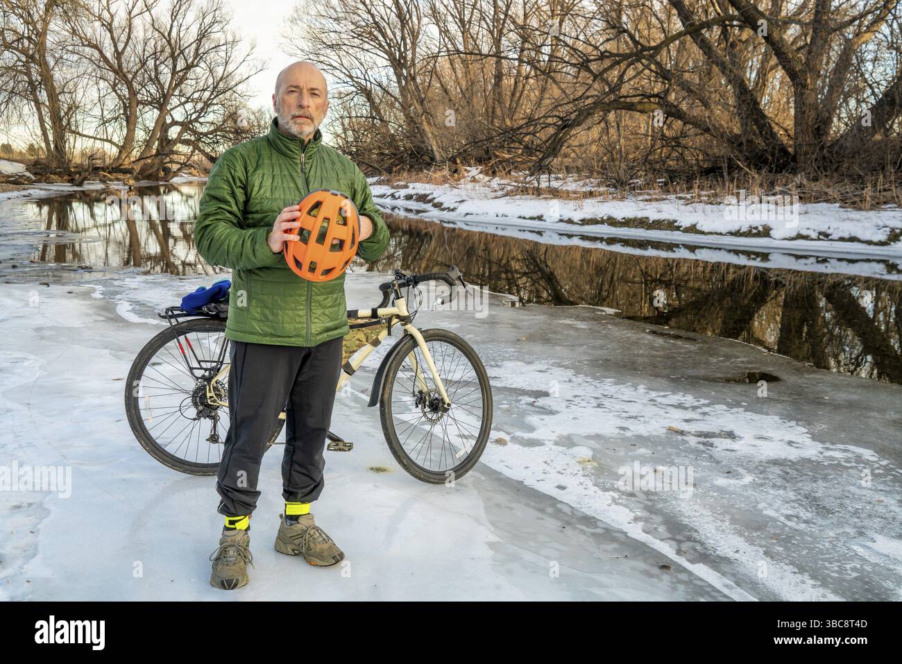 Cycliste masculin senior tenant un casque de protection orange vif est debout avec un vélo de randonnée sur une rive glacée de la rivière, poudre River à Fort Collins, couleur Banque D'Images