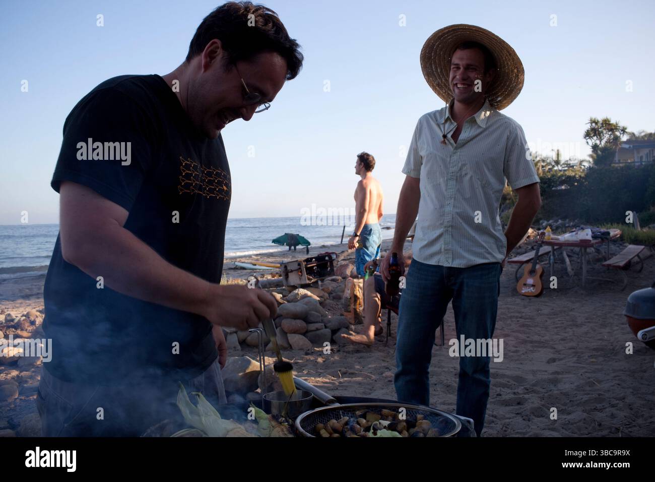 Deux jeunes hommes grillent à la plage tandis qu'un autre regarde le surf au Rincon Beach State Park près de Carpinteria, en Californie. Banque D'Images