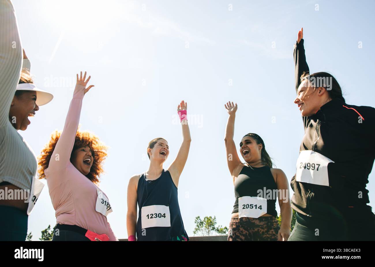 Un groupe diversifié de participants joyeux lèvent les bras pour triompher et célèbrent le travail d'équipe lors d'un événement de course à pied. Ils portent des numéros de course et GE athlétique Banque D'Images