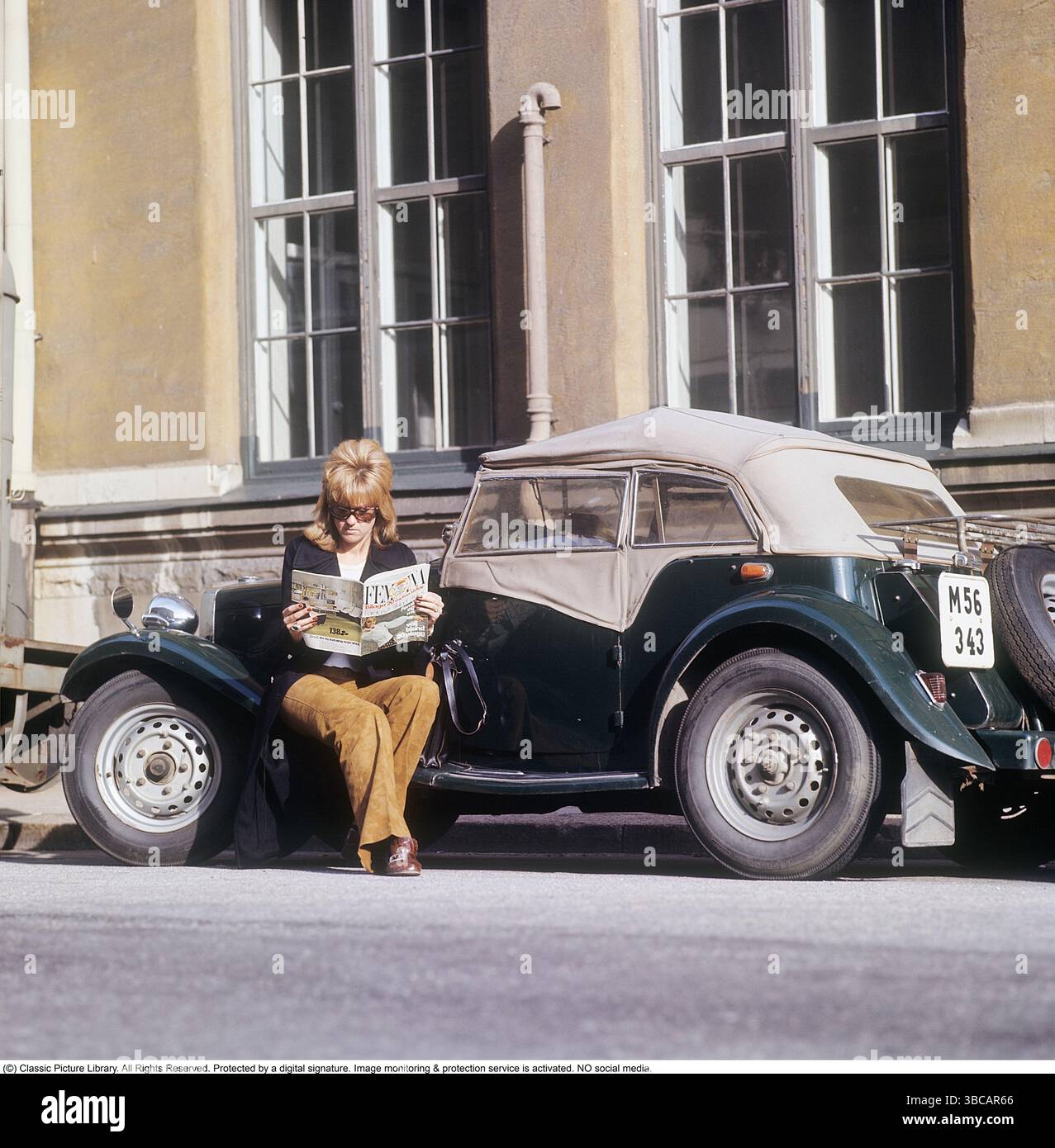 Une femme près d'une voiture de sport. La voiture est un modèle anglais MG TD, produit entre 1950 et 1953. Ce modèle était particulièrement bien adapté aux clients suédois car il pouvait être construit avec le volant sur le côté gauche, aligné sur les conventions de conduite suédoises de l'époque. Au total, 29 664 unités de MG TD ont été fabriquées. Elle est assise sur la voiture, lisant le magazine féminin Femina. Elle est habillée d'un blazer et d'un pantalon marron. Années 1960 Suède. Autres faits sur la MG TD la MG TD, qui fait partie de la série MG T-type, était un roadster biplace réputé pour son design britannique classique, feat Banque D'Images