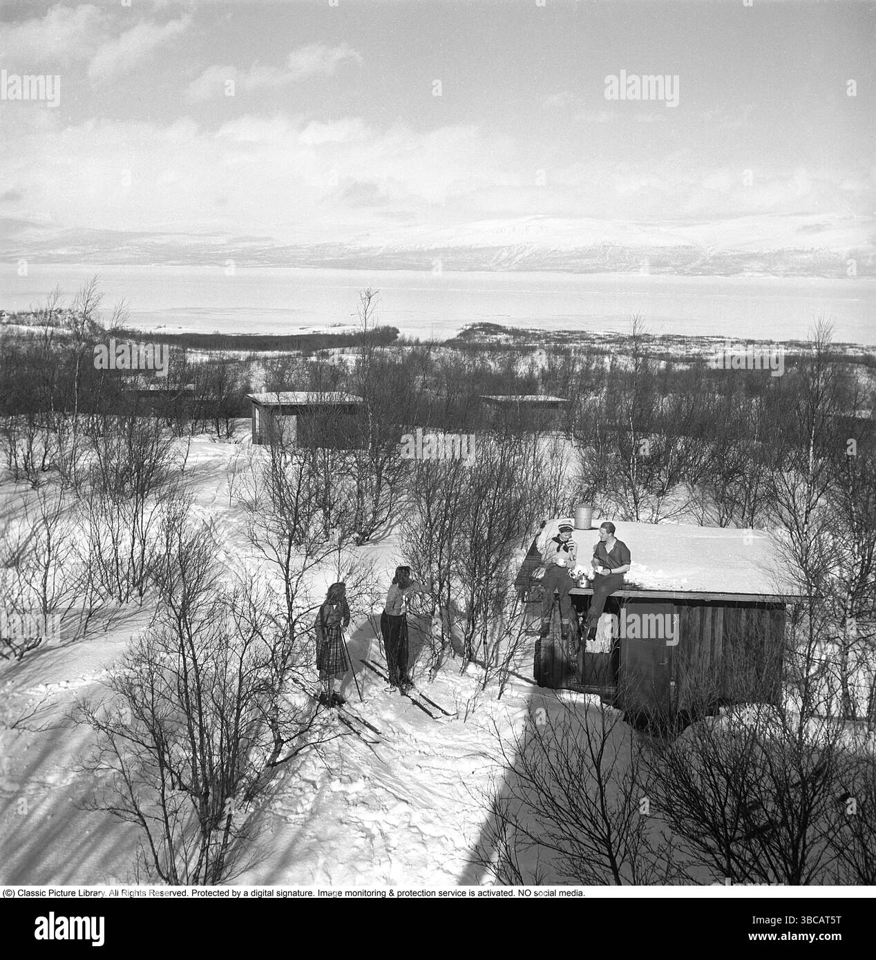 Hiver dans les années 1940 Un jeune couple prend une tasse de café tout en étant assis sur le toit de leur chalet. Leurs deux amis sont debout sur des skis et discutent. La photo est prise en Laponie suédoise près de Björkliden. C'est M. et Mme Iris et Bertil Thorelli qui passent un bon moment dans la nature sauvage suédoise. Suède 1943 photo Kristoffersson Réf. D109-4 Banque D'Images