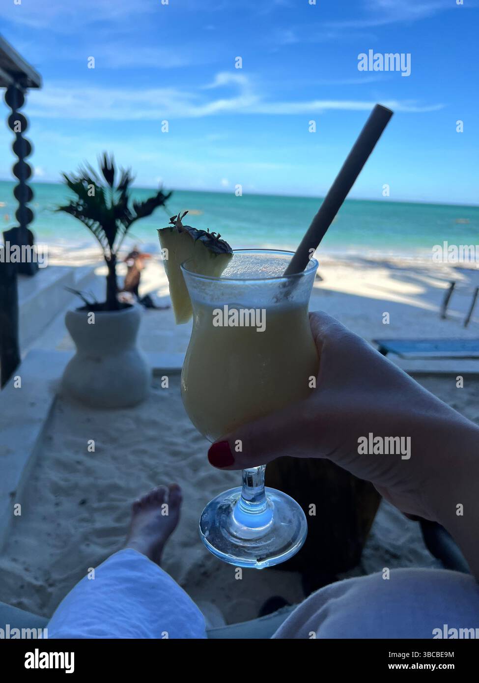 Une piña colada rafraîchissante se tient jusqu’aux eaux turquoises de la côte immaculée de Zanzibar avec les orteils dans le sable blanc doux et un océan Indien chaud. Banque D'Images