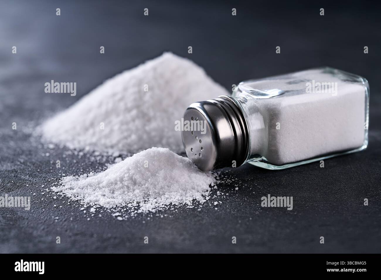 Salière et pile de cristaux de sel renversés sur une table noire, foyer sélectif. Un tas de sel de shaker de sel, concept de consommation excessive de sel. Banque D'Images