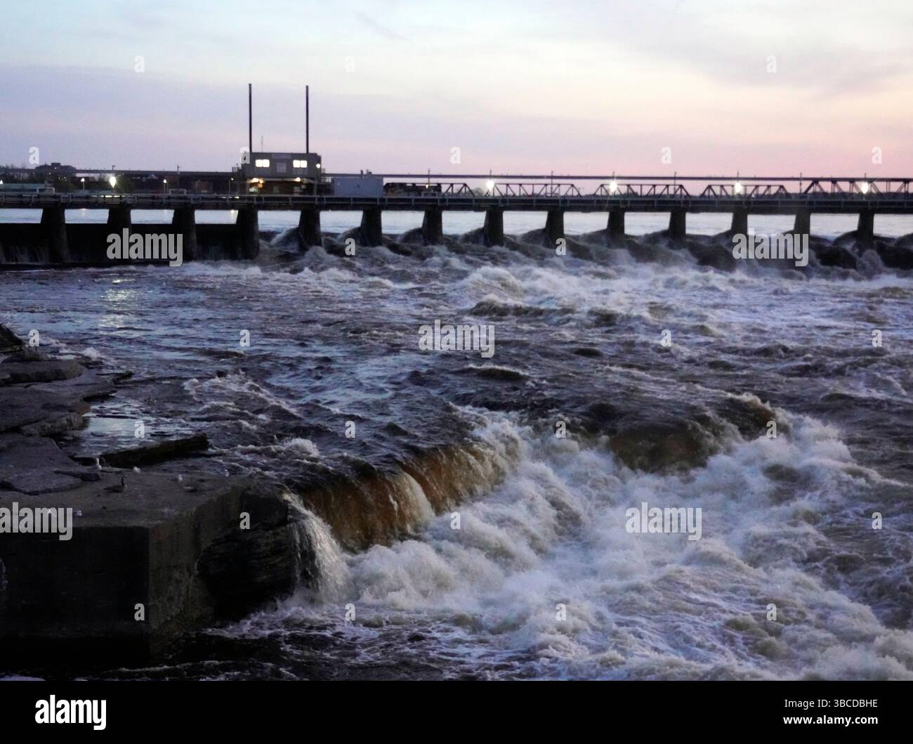 Barrage puissant avec l'eau précipitée enjambe une large rivière au crépuscule, avec des lumières éclairant la structure Banque D'Images