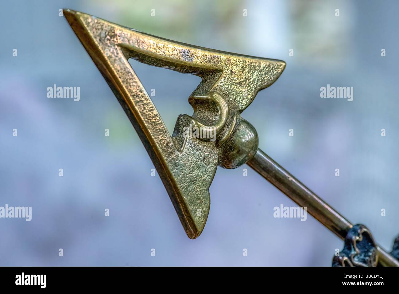 Gros plan d'un ornement en forme de flèche en laiton ou d'un pointeur avec découpe géométrique et texture vieillie, monté sur une tige en métal sur fond doux. Banque D'Images