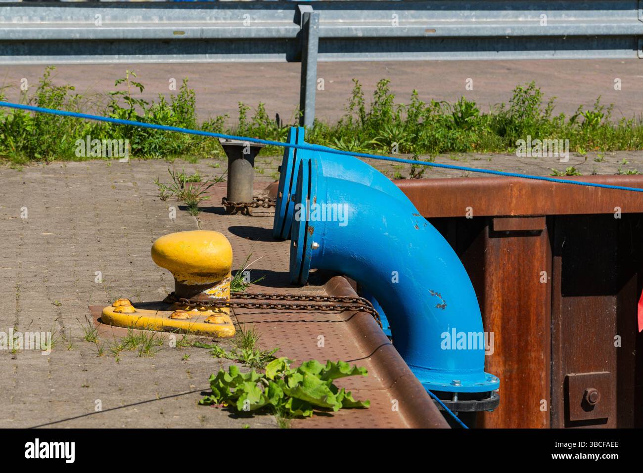 Un tuyau bleu se courbe près d'une borne d'amarrage jaune le long d'un quai. Le quai dispose d'une surface en béton avec des mauvaises herbes poussant, entouré d'un maillon de chaîne fenc Banque D'Images