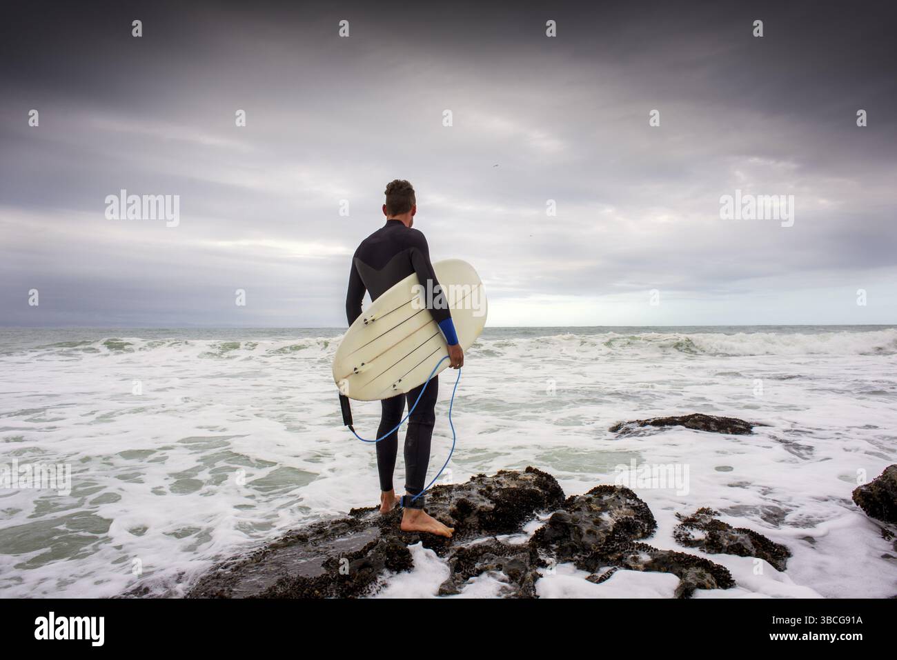 Un surfeur se tient sur les rochers avec sa planche de surf sous le bras alors qu'il regarde le surf juste avant de sauter dans l'eau Banque D'Images