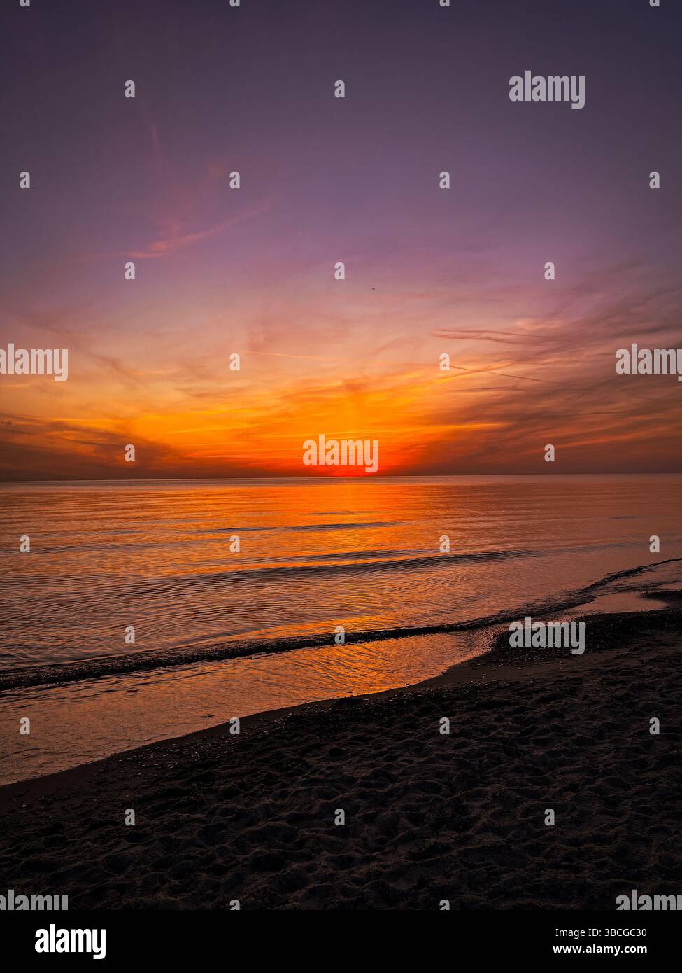 Coucher de soleil spectaculaire sur la mer Baltique. Ciel orange et violet éclatant se reflétant sur l'eau, avec des vagues douces roulant sur le rivage sablonneux. Banque D'Images