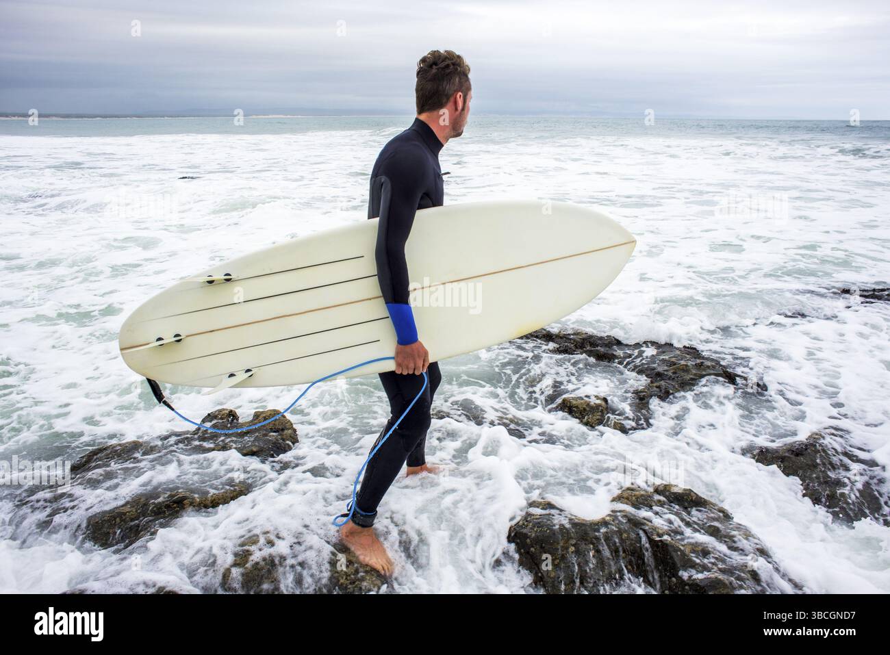Un homme marche sur les rochers tandis que la vague roule dessus tandis qu'il tient une planche de surf dans son bras Banque D'Images