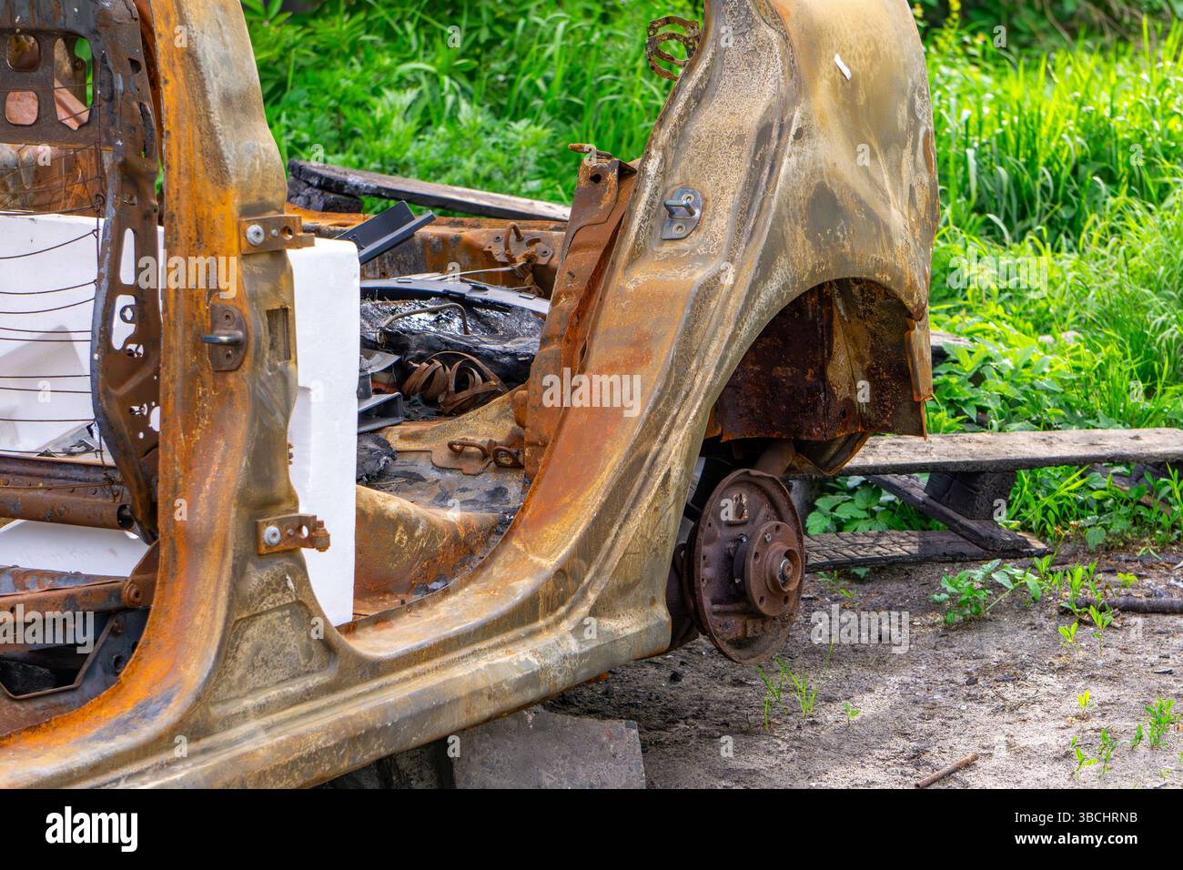 Coquille rouillée d'une voiture brûlée sans portes ni roues, placée sur le sol entourée d'herbe. Banque D'Images