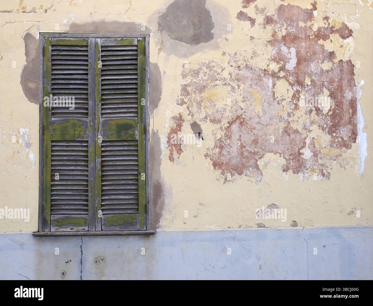 Un vieux mur de maison fissuré avec du ciment patché peint dans des couches écaillées de peinture grise jaune et rouge avec une fenêtre en bois fissuré vert avec c Banque D'Images
