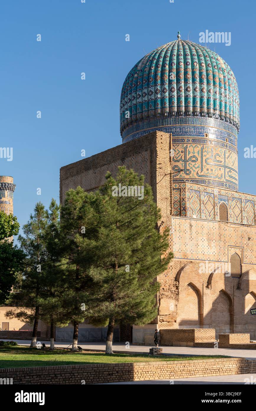 Un massif dôme rond turquoise se trouve au sommet de solides murs de briques sur le côté nord de la mosquée Bibi-Khanym à Samarcande, Ouzbékistan, parmi les arbres verts. Banque D'Images