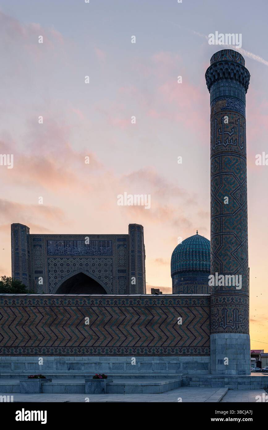 Le dôme et l’entrée de la mosquée Bibi-Khanym brillent au coucher du soleil à Samarcande, en Ouzbékistan, avec des arches en briques et un minaret formant une silhouette saisissante. Banque D'Images