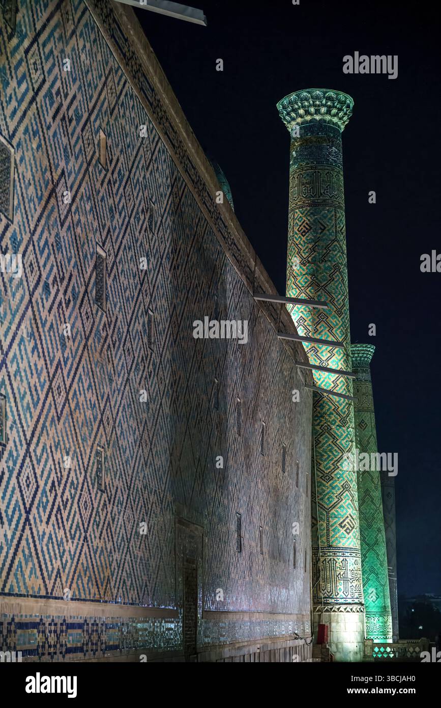 Une vue nocturne de Sherdor Madrasa au Registan à Samarcande, Ouzbékistan, avec un grand minaret et des murs de briques à motifs illuminés sous le ciel sombre. Banque D'Images