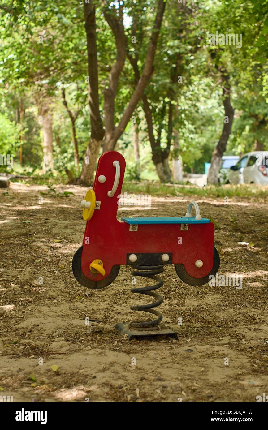 Un rocker à ressort rouge vif en forme de moto attend les petits coureurs sur un terrain de jeu ombragé par une journée ensoleillée. Banque D'Images