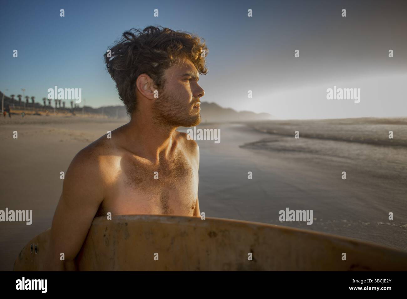 Portrait d'un jeune surfeur, avec une planche de surf sous le bras, debout sur la plage tout en inspectant les vagues dans la lumière du soleil tôt le matin Banque D'Images