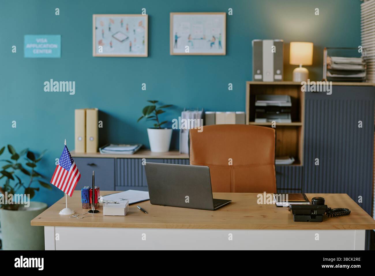 Centre visa avec bureau en bois avec fournitures organisées, ordinateur portable et drapeau américain. Mur orné d'œuvres d'art colorées et lampe éclairée, fournissant produ Banque D'Images