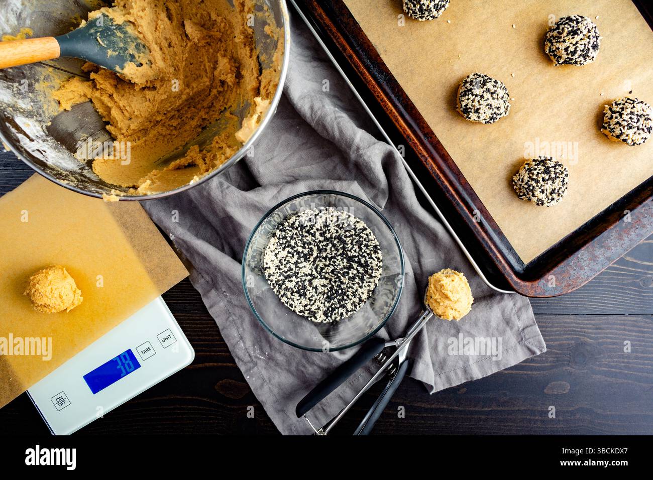Boules de pâte à biscuits roulées dans des graines de sésame : pâte à biscuits, graines de sésame blanches et noires, cuillère à biscuits, balance de cuisine et poêle en feuille parchemin Banque D'Images
