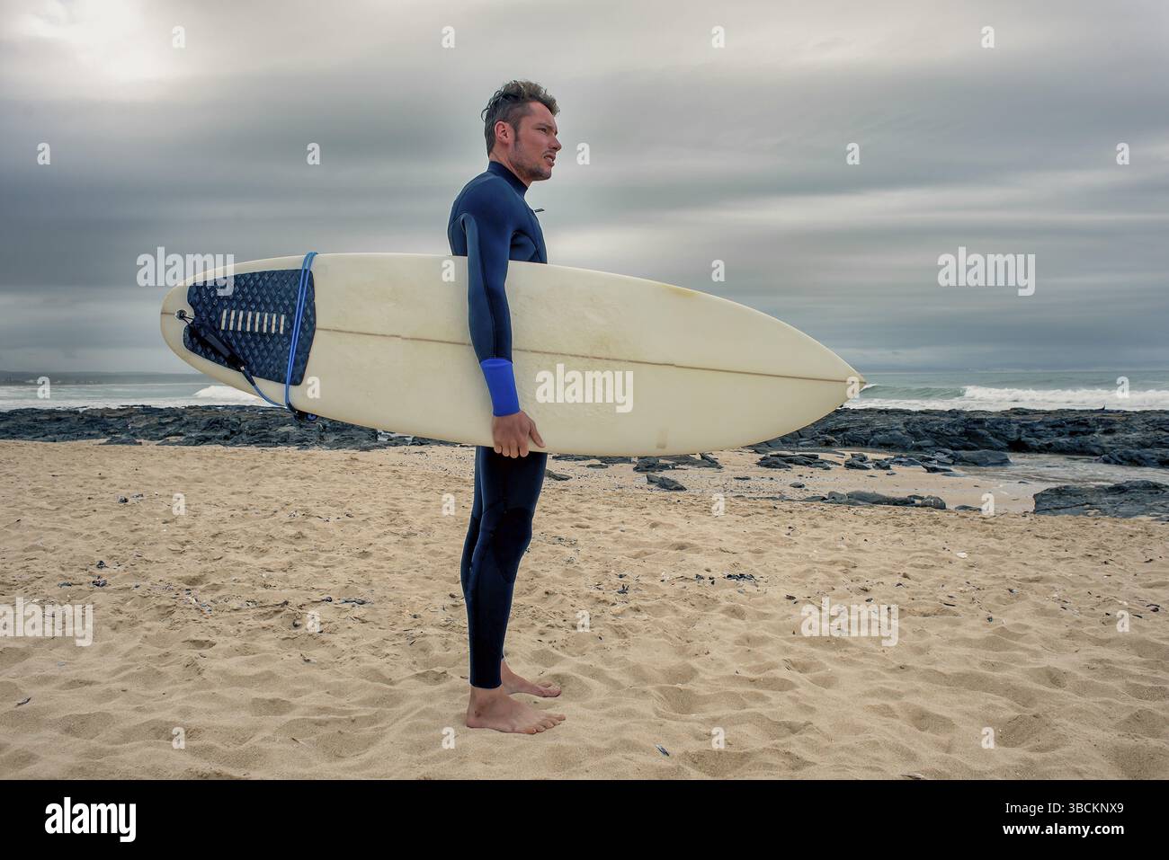 Un portrait en longueur du côté d'un surfeur debout sur la plage avec une planche de surf sous le bras alors qu'il regarde vers l'océan Banque D'Images
