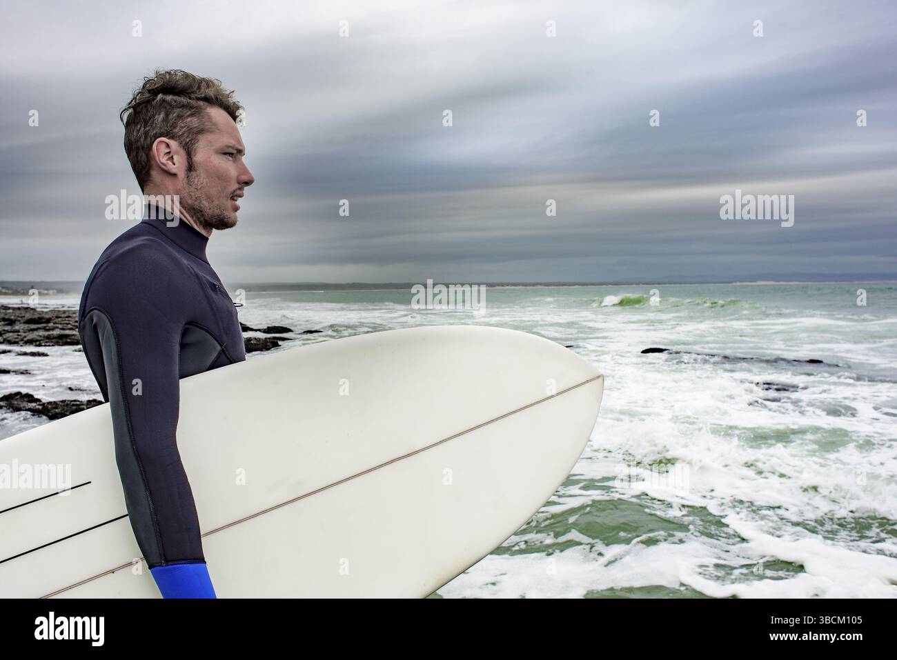 Portrait de côté d'un surfeur, avec sa planche de surf sous le bras alors qu'il regarde l'océan Banque D'Images