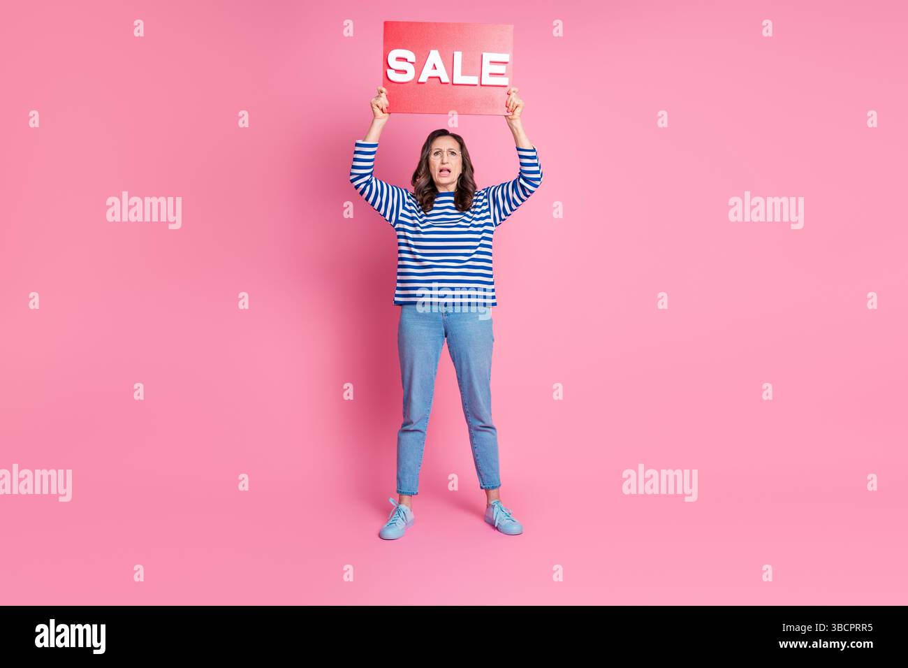 Femme d'âge moyen à la mode tenant un signe de vente rouge dans la pose debout sur fond rose vif représentant la vente au détail Banque D'Images
