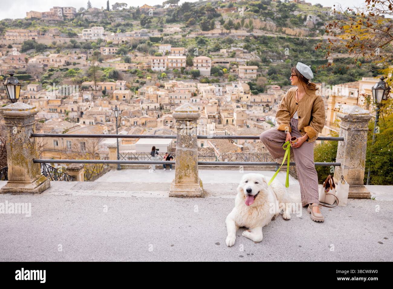 Femme avec grand chien blanc profite de la vue panoramique sur Modica, Sicile. Un moment parfait de connexion, de sérénité et de lenteur de voyage entouré de toits en terre cuite et d'architecture baroque Banque D'Images