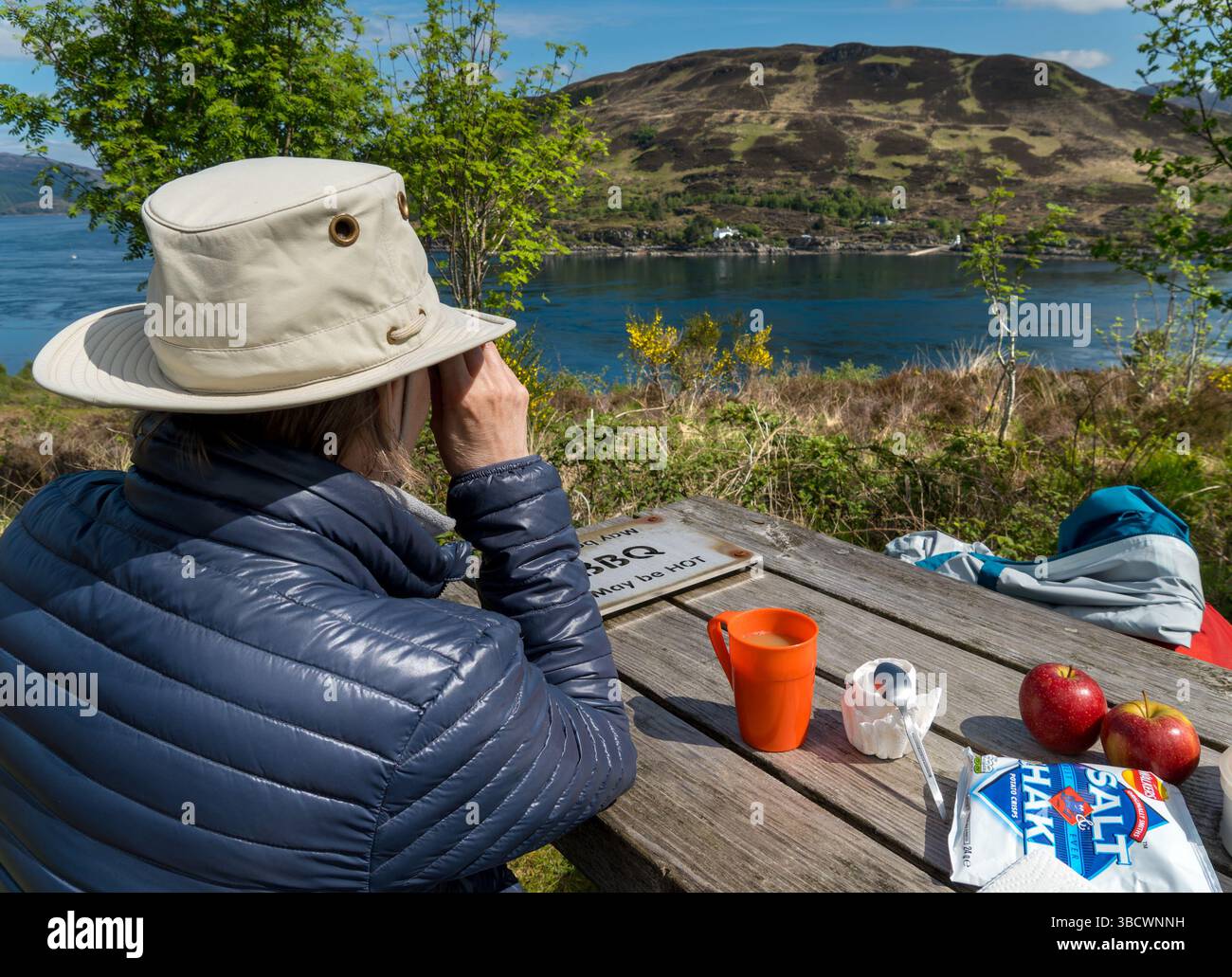 Femme en chapeau de soleil et blouson gonflant regardant la vue de la table de pique-nique Kylerhea sur l'île de Skye au-dessus de Kyle Rhea sur le continent écossais à Glenelg. Banque D'Images