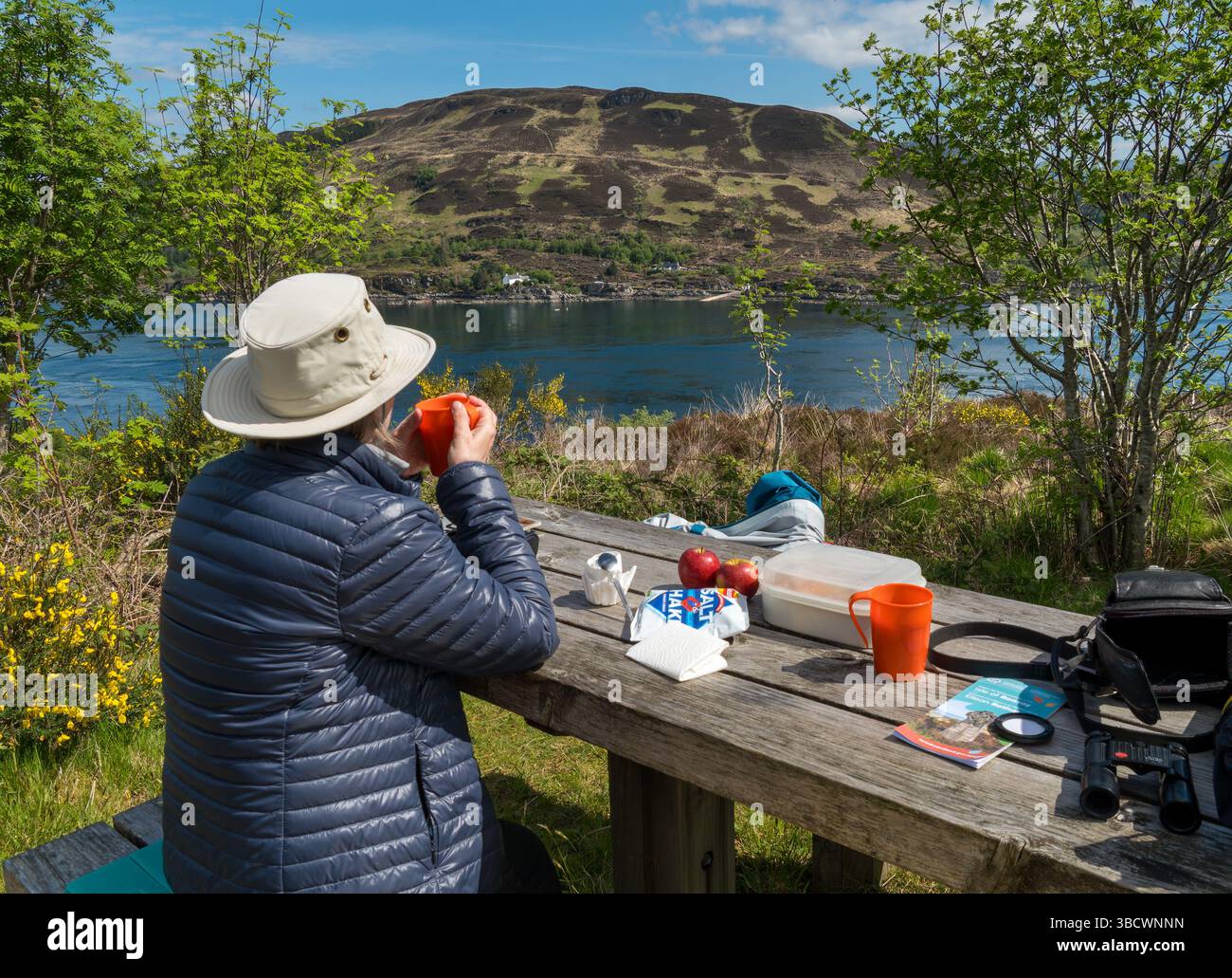 Femme en chapeau de soleil et blouson gonflant regardant la vue de la table de pique-nique Kylerhea sur l'île de Skye au-dessus de Kyle Rhea sur le continent écossais à Glenelg. Banque D'Images