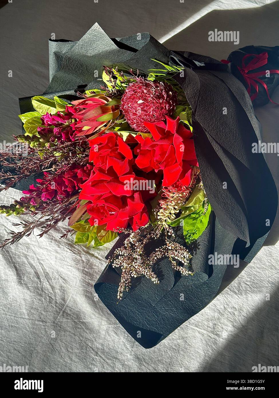 Élégant bouquet de fleurs rouges vibrantes avec de la verdure, enveloppé dans du papier noir et affiché à la lumière naturelle du soleil sur une surface de tissu blanc - Image de stock capturée avec un smartphone