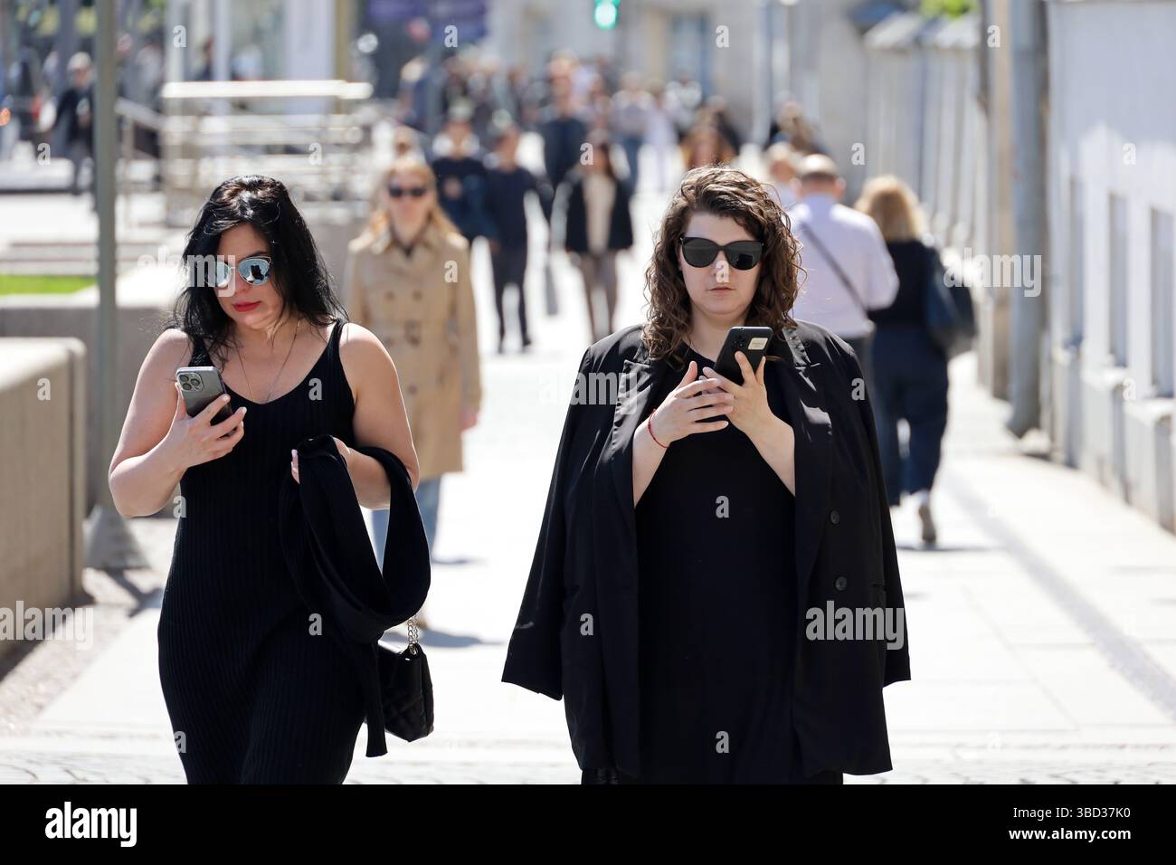 Foule de gens marchant dans une rue de la ville, deux femmes en lunettes de soleil utilisant des smartphones au premier plan Banque D'Images
