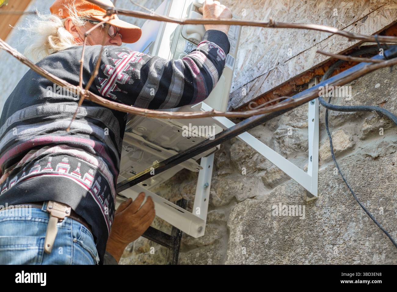 Un homme installe un climatiseur extérieur sur des supports de montage en fer sur le mur d'une maison rurale en pierre. Installation DIY. Banque D'Images