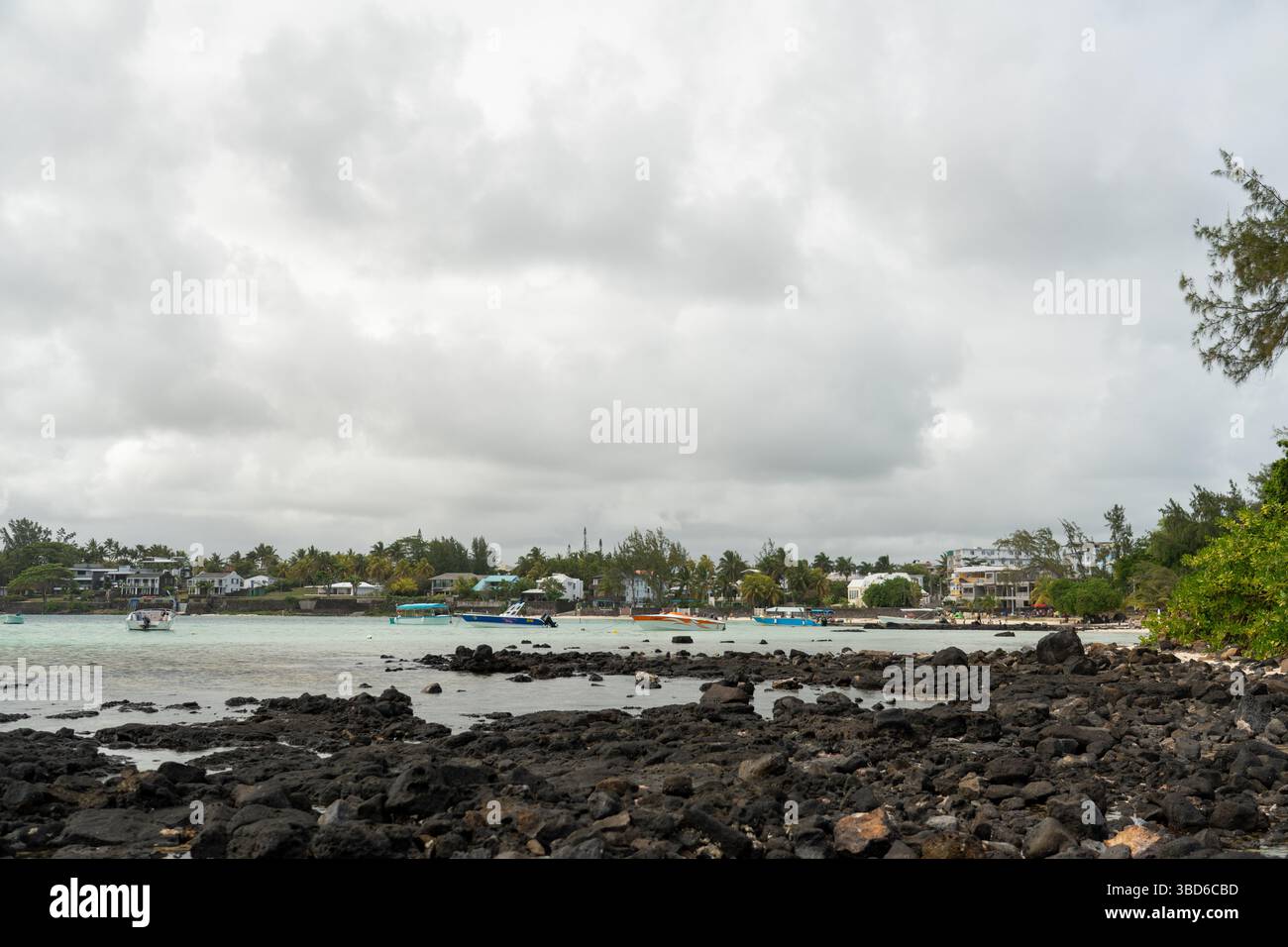 Le Morne, Maurice - 12 mai 2024 : vue panoramique sur le rivage rocheux avec des bateaux ancrés, village et végétation luxuriante sous ciel nuageux Banque D'Images