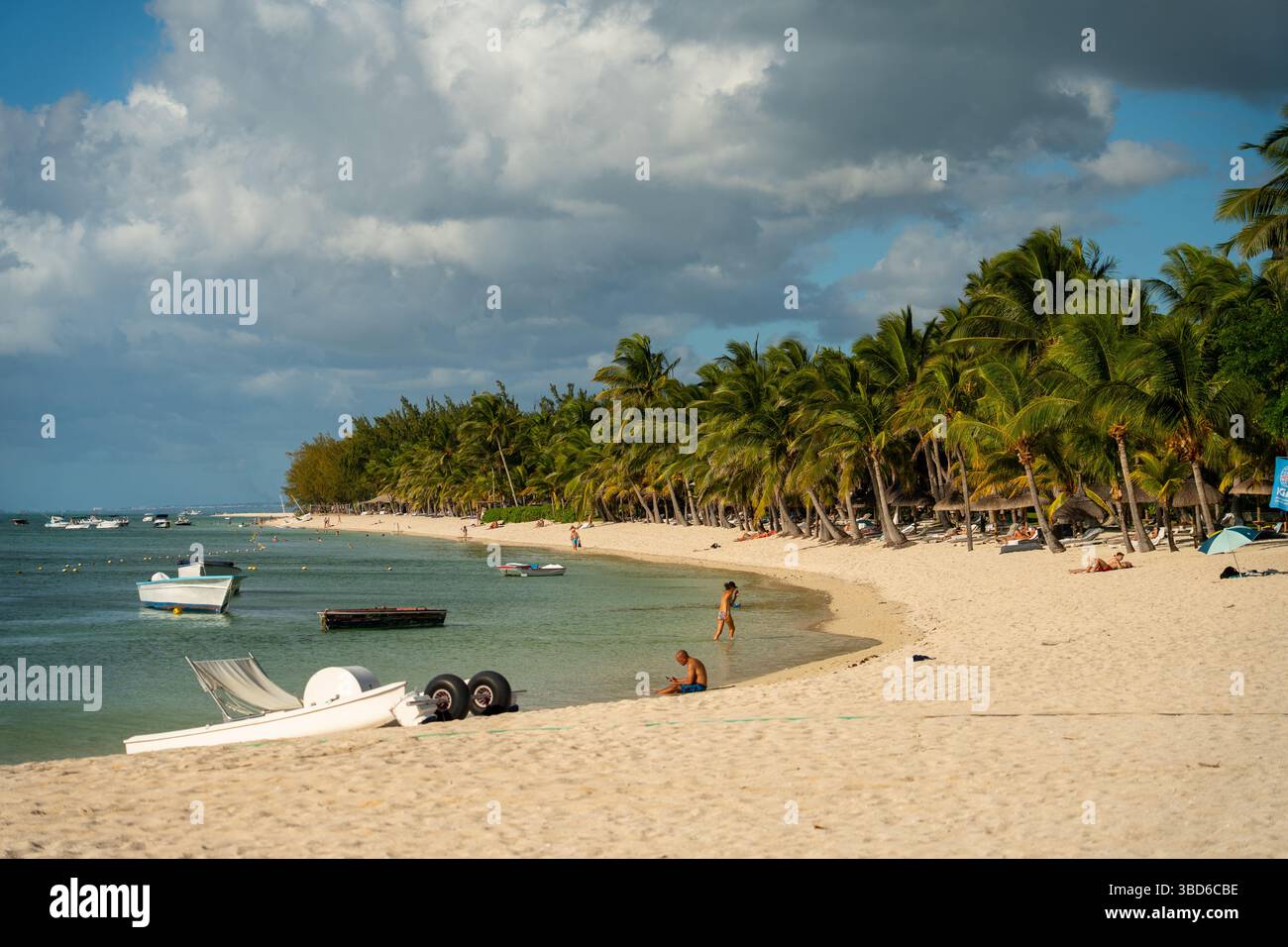 Le Morne, Maurice - 12 mai 2024 : les gens marchent et se détendent sur une longue plage de sable bordée de palmiers et de petits bateaux ancrés au large Banque D'Images