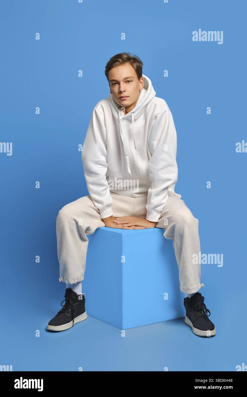 Homme gai avec un sweat-shirt blanc et un pantalon de survêtement assis sur un cube en bois sur fond bleu de studio Banque D'Images