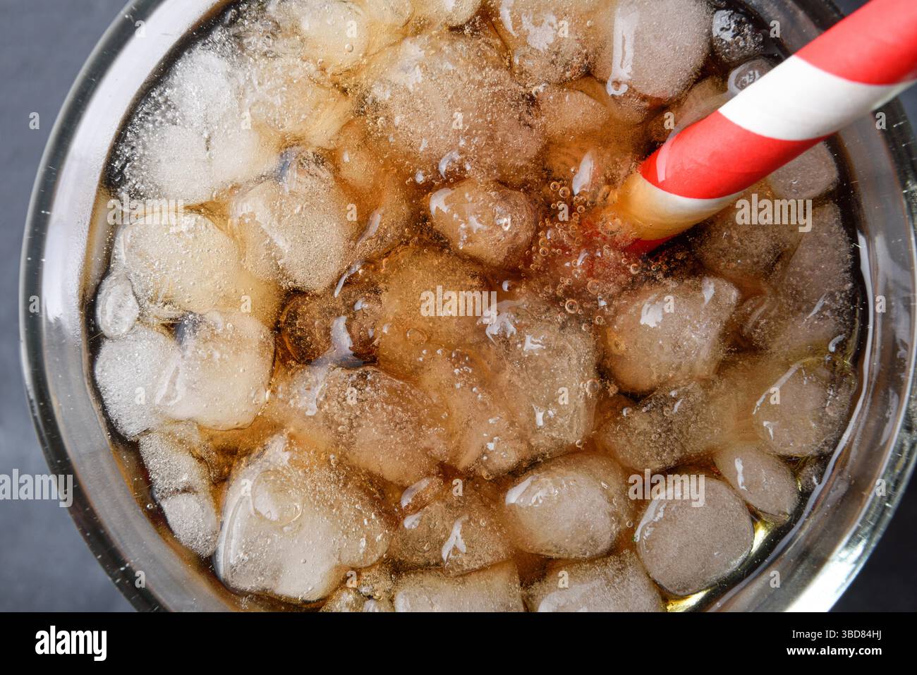 Détail de la bouche d'un verre transparent avec une boisson rafraîchissante avec de la glace et une paille blanche et rouge sur un fond sombre. Vue de dessus. Banque D'Images