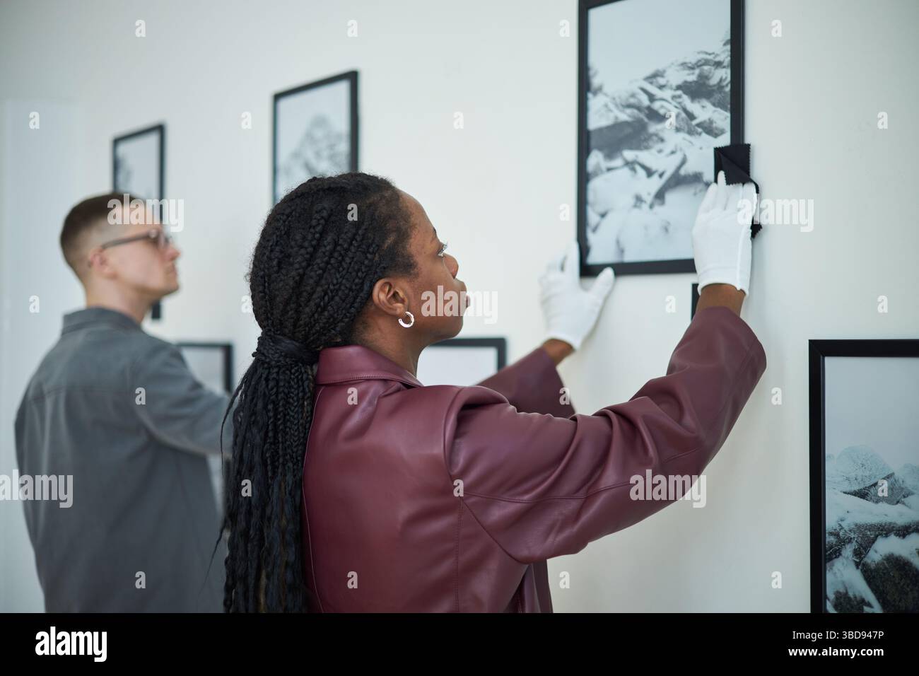 Femme et homme accrochant des œuvres d'art encadrées sur le mur blanc dans la galerie d'art, portant des gants de travail et positionnant soigneusement les pièces. Travail d'équipe et attention à de Banque D'Images