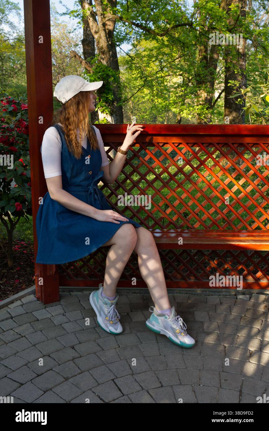 Une fille dans une robe bleue et un chapeau blanc est assis sur un banc en bois dans un jardin luxuriant, entouré de fleurs et d'arbres, profitant de l'atmosphère paisible en plein air Banque D'Images