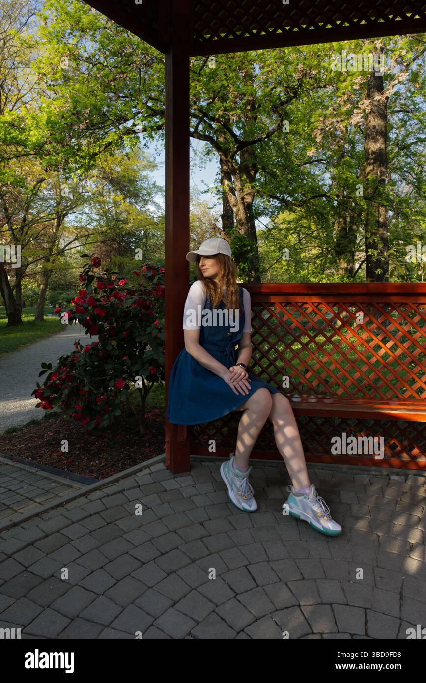 Une fille dans une robe bleue et un chapeau blanc est assis sur un banc en bois dans un jardin luxuriant, entouré de fleurs et d'arbres, profitant de l'atmosphère paisible en plein air Banque D'Images
