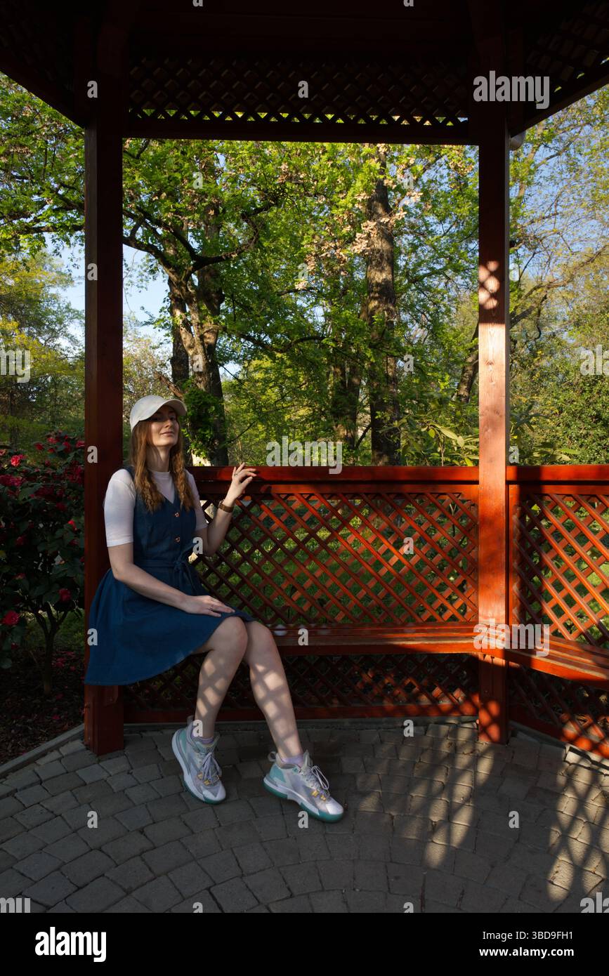 Une fille dans une robe bleue et un chapeau blanc est assis sur un banc en bois dans un jardin luxuriant, entouré de fleurs et d'arbres, profitant de l'atmosphère paisible en plein air Banque D'Images