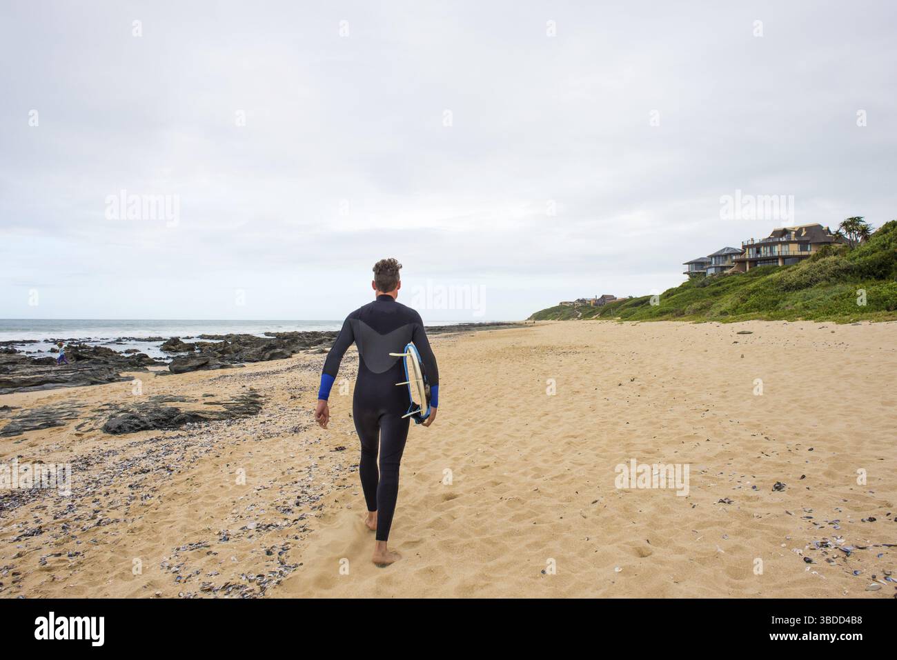 Un surfeur en combinaison marche le long de la plage avec sa planche de surf sous le bras Banque D'Images