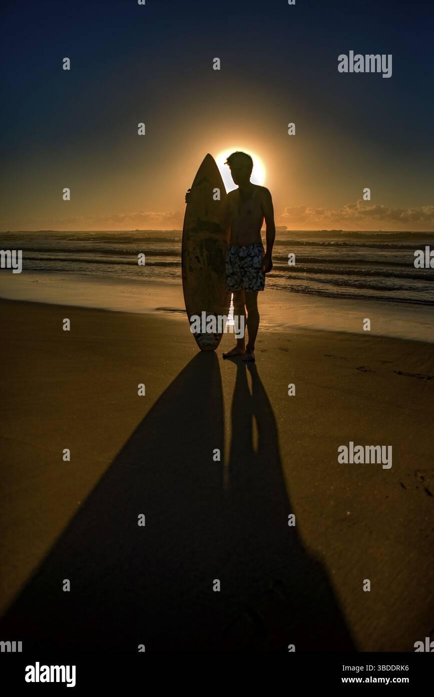Un jeune surfeur se tient debout avec une planche de surf à la main sur la plage juste en face du soleil levant créant une longue ombre devant le surfeur Banque D'Images