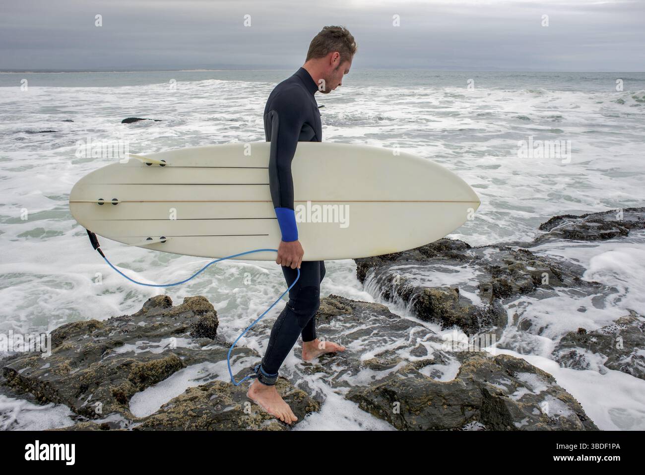Un surfeur avec sa planche de surf sous le bras marche sur quelques rochers alors qu'il approche de l'océan pour aller faire un surf Banque D'Images