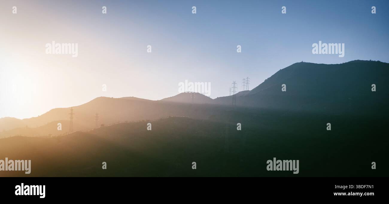 Coucher de soleil sur des collines vallonnées avec des silhouettes de pylônes électriques, signifiant un mélange de nature et d'infrastructure humaine dans un cadre tranquille Banque D'Images