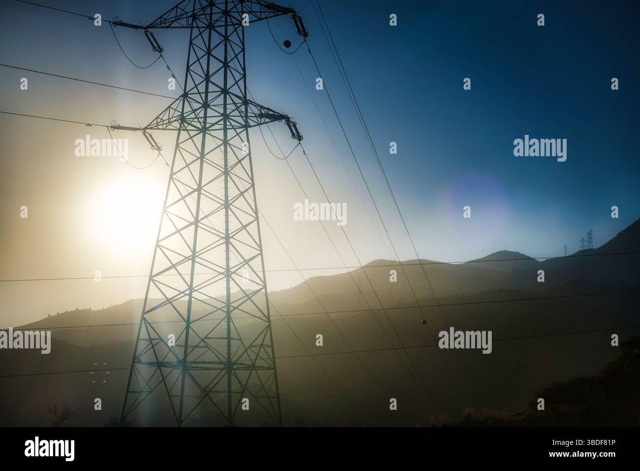 Image dramatique d'une tour de transmission de puissance silhouette contre un ciel brumeux éclairé par le soleil, avec des collines en arrière-plan partiellement enveloppées de brume Banque D'Images