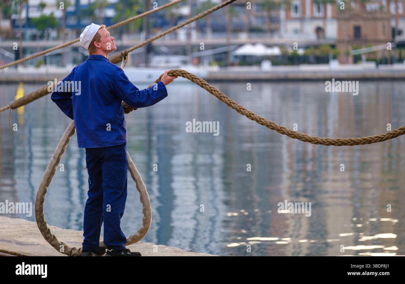 Espagne Malaga 1 28 2024 Sailor en uniforme bleu saisissant fermement une corde épaisse au port urbain, indicatif des activités maritimes Banque D'Images
